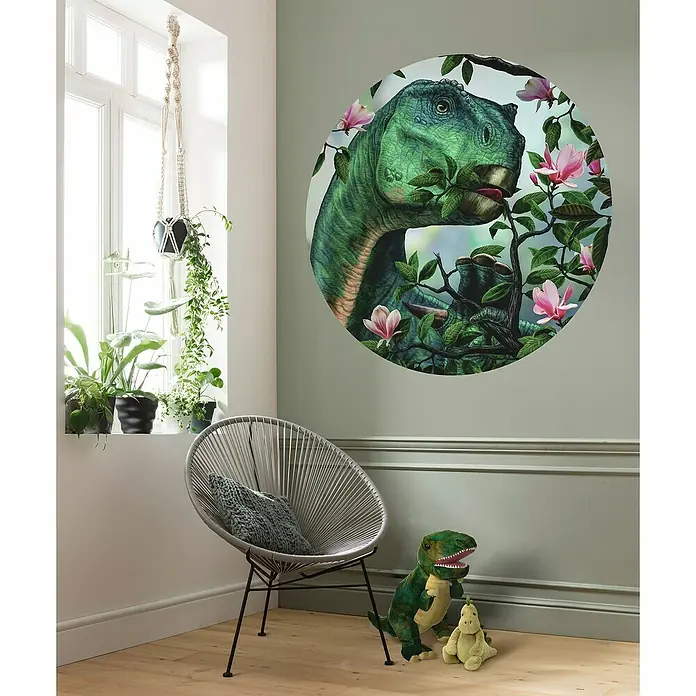 Komar Fototapete rund Iguanodon Flowers - 128 cm, Selbstklebend - Ein Kinderzimmer mit einem Dinosaurier-Wandaufkleber, einem grauen Stuhl und einer Fensterbank mit Pflanzen.