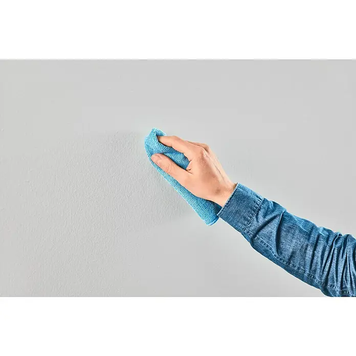 Tesa Powerstrips Klebehaken Adjust Keyhole - Kunststoff, Weiß - Nahaufnahme einer Person, die eine graue Wand mit einem blauen Mikrofaser-Reinigungstuch reinigt, um Staub und Schmutz zu entfernen.