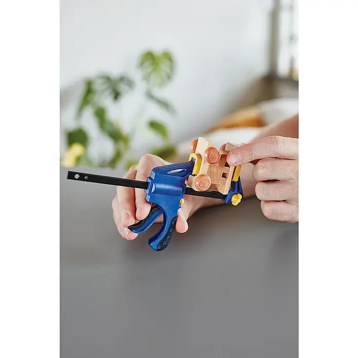 Rapid Einhandzwinge - Spannweite: 100 mm - Nahaufnahme einer blauen Stangenzwinge, die ein Holzspielzeugauto auf einer grauen Oberfläche hält, im Hintergrund eine Pflanze.