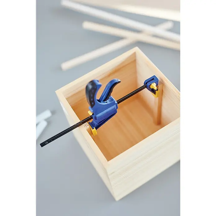 Rapid Einhandzwinge - Spannweite: 100 mm - Holzkiste wird mit einer blauen Klemme fixiert, um Stabilität beim Basteln zu gewährleisten, während Holzstäbe im Hintergrund liegen.