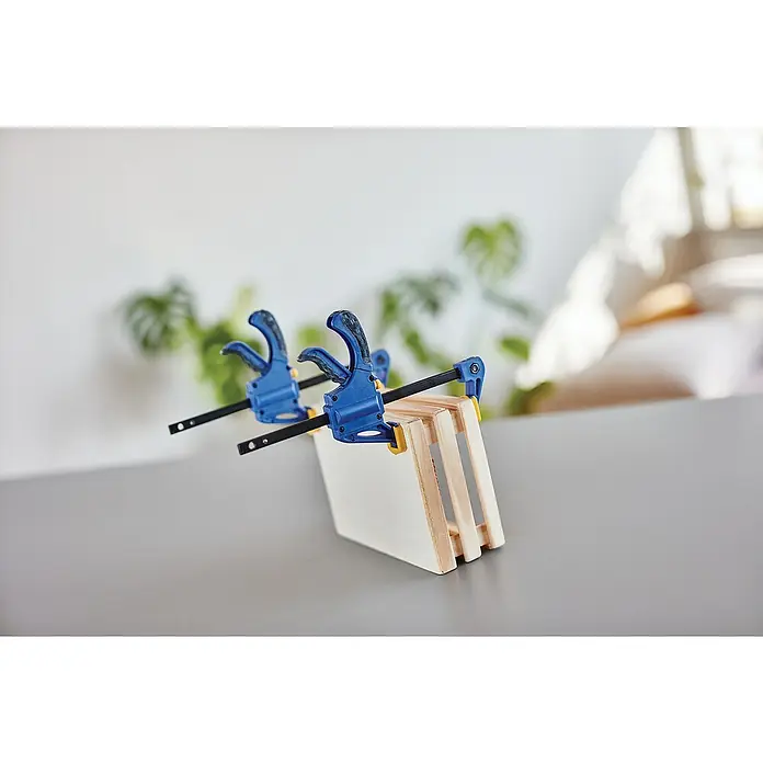 Rapid Einhandzwinge - Spannweite: 100 mm - Zwei blaue Federklemmen fixieren ein Holzprojekt auf einem grauen Tisch, im Hintergrund eine Pflanze, wodurch der Fokus auf das Werkzeug und die Holzbearbeitung gelegt wird.