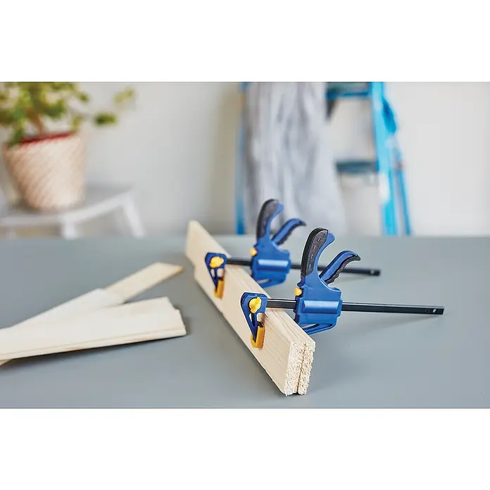 Rapid Einhandzwinge - Spannweite: 100 mm - Zwei blaue Klemmen halten Holzstücke auf einer grauen Oberfläche zusammen, mit einer Leiter und einer Pflanze im Hintergrund.