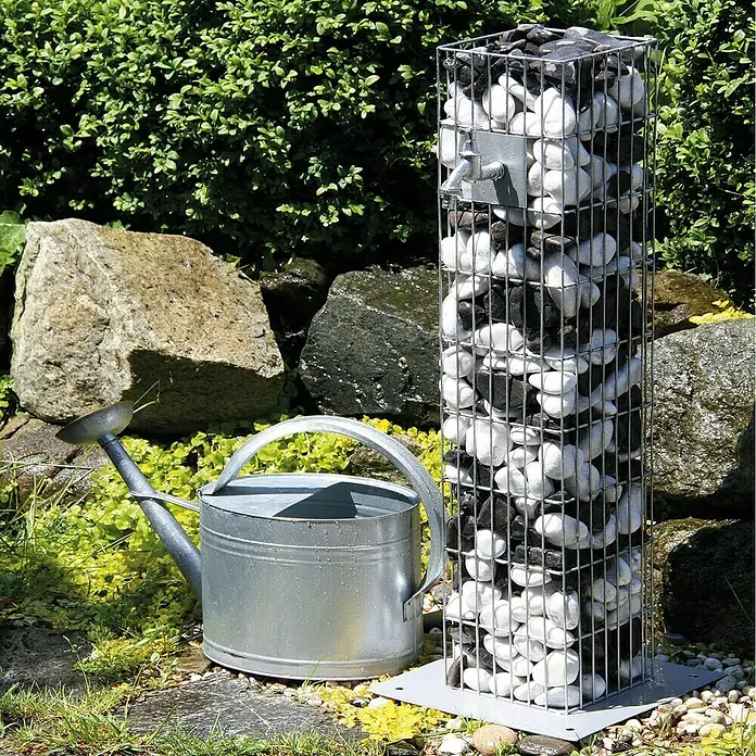 Bellissa Gabionensäule Gabione18,5 x 18,5 x 90,5 cm, Stahl, Maschenbreite: 25 mm, Maschenhöhe: 100 mm - Gartenszene mit einer stilvollen Gabione, gefüllt mit schwarzen und weißen Kieselsteinen, neben einer metallenen Gießkanne.