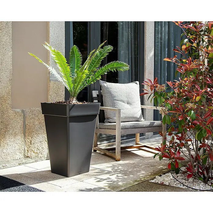 Artevasi Capri Pflanztopf - Außenmaß (L x B x H): 30 x 30 x 49,5 cm, Kunststoff, Anthrazit - Ein hoher, rechteckiger, schwarzer Pflanzkasten mit einer Sago-Palme darauf, steht neben einem Stuhl im Freien.