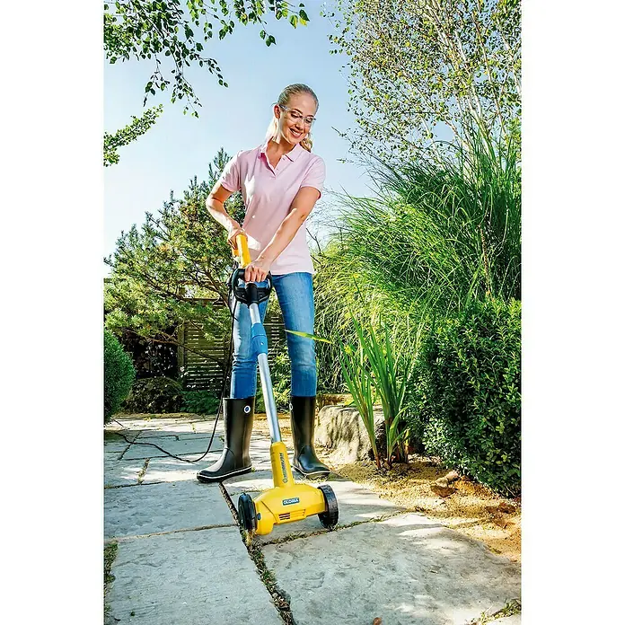 Gloria Power for All 18V Fugenreiniger WeedBrush li-on - 18 V, Li-Ionen, Ohne Akku - Eine Frau benutzt einen Gloria Weedbrush zur Unkrautentfernung auf einer Steinterrasse, umgeben von üppigem Grün.