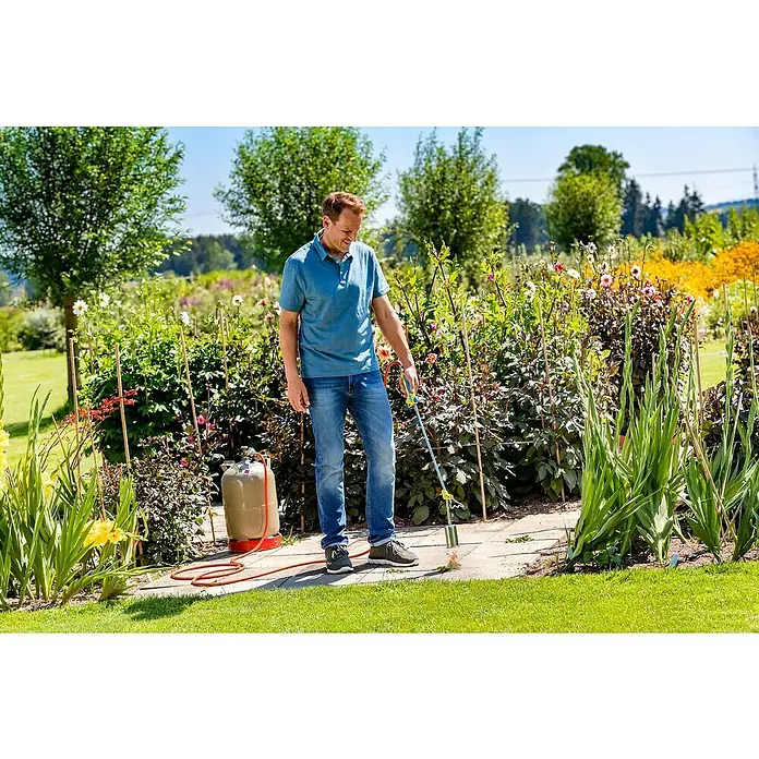 Gloria Unkrautvernichter Thermoflamm bio Professional PLUS - Flammtemperatur: 1.000 °C - Ein Mann verwendet einen Abflammgerät, um Unkraut von einem Gehweg in einem üppigen Garten zu entfernen, mit einer Propangasflasche in der Nähe.