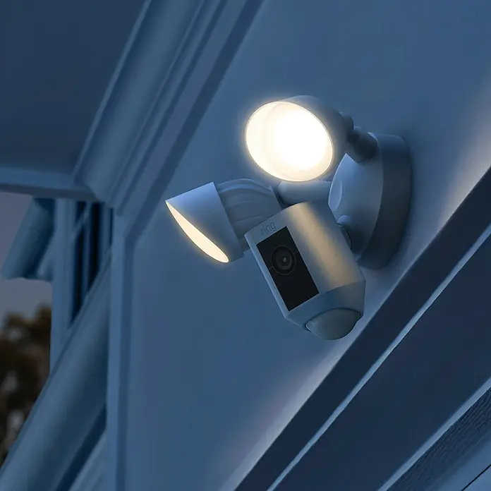 Ring Überwachungskamera Flutlicht Plus Kabel - Netzbetrieben, 1.920 x 1.080 Pixel (Full HD), Weiß - Nahaufnahme einer Ring Floodlight Cam, die an einer Wand montiert ist, mit zwei hell leuchtenden Scheinwerfern für mehr Sicherheit.