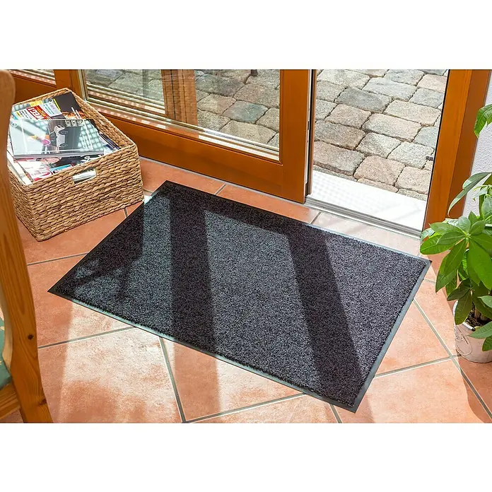 Astra Schmutzfangmatte Proper Tex - Schwarz, 60 x 180 cm, Material Nutzschicht: 100 % Polyamid - Eine graue Fußmatte liegt vor einer offenen Tür auf einem gefliesten Boden, neben einem Korb mit Zeitschriften und einer Topfpflanze.