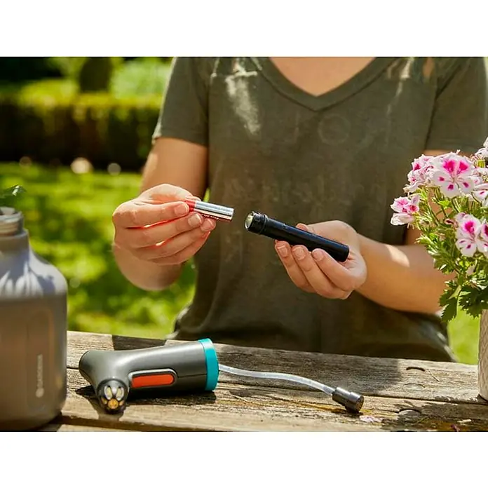 Gardena Handsprüher Easy Pump1 l - Eine Person legt Batterien in eine GARDENA Akku-Druckspritze, die auf einem Holztisch liegt, daneben ein Behälter und eine blühende Pflanze.