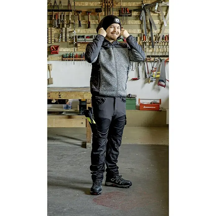 L.Brador Arbeitshose 1090PB - 50, Schwarz - Ein Mann in Arbeitskleidung, inklusive Jacke und Hose, mit einer Stirnlampe auf dem Kopf, vor einer Werkzeugwand.