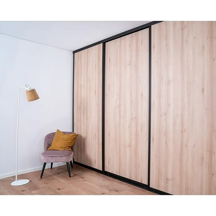 Room Plaza Easy Doing Schiebetür-Bau-Set - Buche Naturwuchs, Farbe Beschlag: Schwarz - Ein moderner Schrank mit Schiebetüren in Holzoptik und schwarzen Rahmen steht neben einem Sessel mit Kissen und einer Stehlampe.