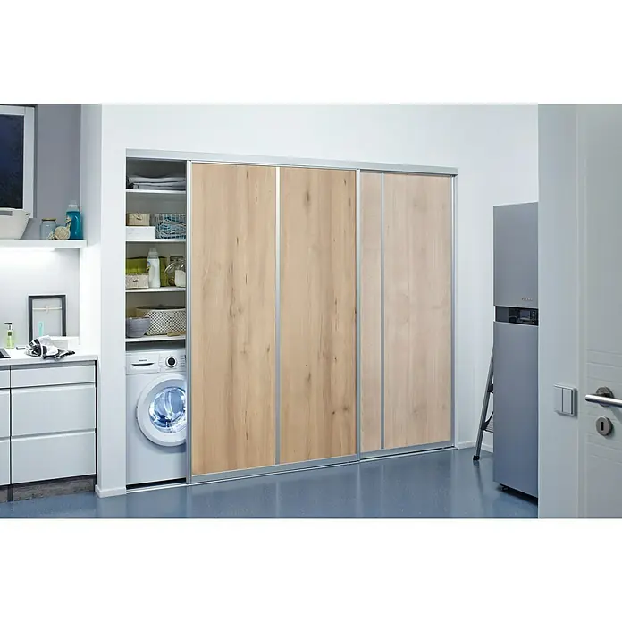 Room Plaza Easy Schiebetür-Bau-Set - Buche Naturwuchs/Ahorn Naturwuchs, Farbe Beschlag: Silber - Eine Waschküche mit einem weißen Waschbecken, Regalen und einer Waschmaschine, die hinter einer Schiebetür mit Holzstruktur versteckt ist.