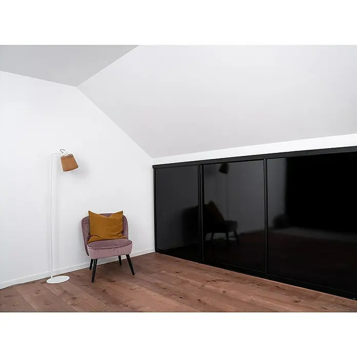 Room Plaza Easy Doing Schiebetür-Bau-Set Kniestock - Hochglanz Schwarz/Hochglanz Weiß, Farbe Beschlag: Schwarz - Innenaufnahme eines Zimmers mit einem schlichten schwarzen Schrank mit Schiebetüren, einem Sessel und einer Stehlampe vor einer weißen Wand.