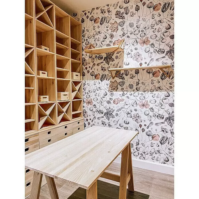 Astigarraga Dinamic Holzregal - H x B x T: 174,6 x 36,2 x 33 cm, Traglast pro Boden: 50 kg, Anzahl Böden: 5 Stk., Schrauben, Natur - Helles Holzregal mit Fächern, X-Unterteilungen und Schubladen, plus ein passender Holztisch in einem Raum mit gemusterter Tapete.