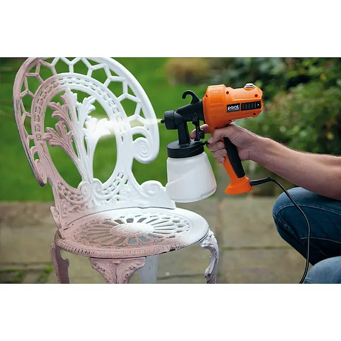 Sistema de pulverización de pintura Paint Sprayer - 400 W, Caudal: 0,5 l/min - Una persona utiliza un pulverizador de pintura Point para aplicar una capa de pintura blanca a una silla de jardín de metal ornamentada al aire libre.