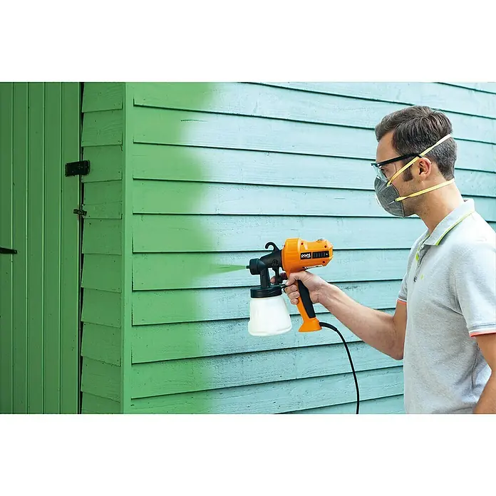 Sistema de pulverización de pintura Paint Sprayer - 400 W, Caudal: 0,5 l/min - Un hombre está usando un rociador de pintura naranja en una pared de madera azul mientras usa una máscara y gafas protectoras.