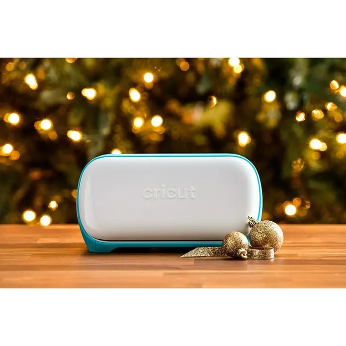 Cricut Joy Schneideplotter Smarte Schneide- und Zeichenmaschine - Türkis/Weiß, Bluetooth, Schnittbreite: 10,4 cm, Schnittlänge: 1,2 m - Nahaufnahme eines weißen Cricut Joy Schneideplotters mit blauem Akzent, platziert auf einem Tisch mit goldenen Weihnachtskugeln und einer Lichterkette im Hintergrund.