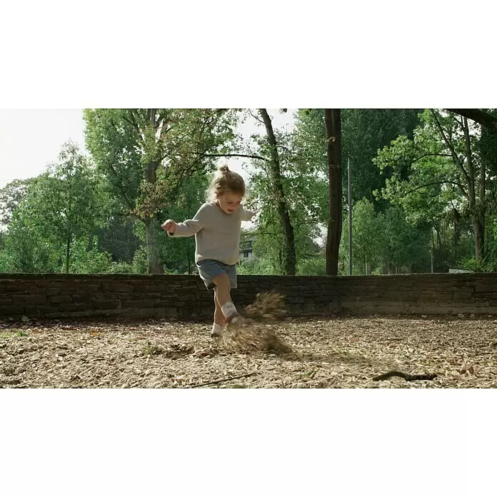 Gardol Pure Nature Hochbeet-Grundfüllung - 50 l, Natur - Ein Kind in hellem Pullover und Jeans-Shorts tritt fröhlich Holzspäne in einem Park.