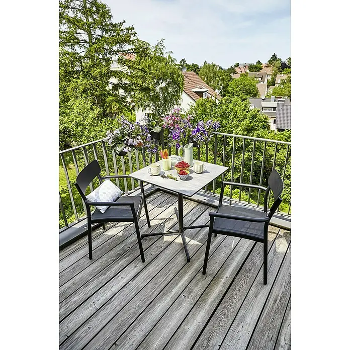 Sunfun Melina Balkontisch - L x B x H: 70 x 70 x 73 cm, HPL, Stahl, Klappbar, Quadratisch, Schwarz - Ein quadratischer Tisch und zwei schwarze Stühle stehen auf einem Balkon mit Holzboden, umgeben von Pflanzen und Blick ins Grüne.