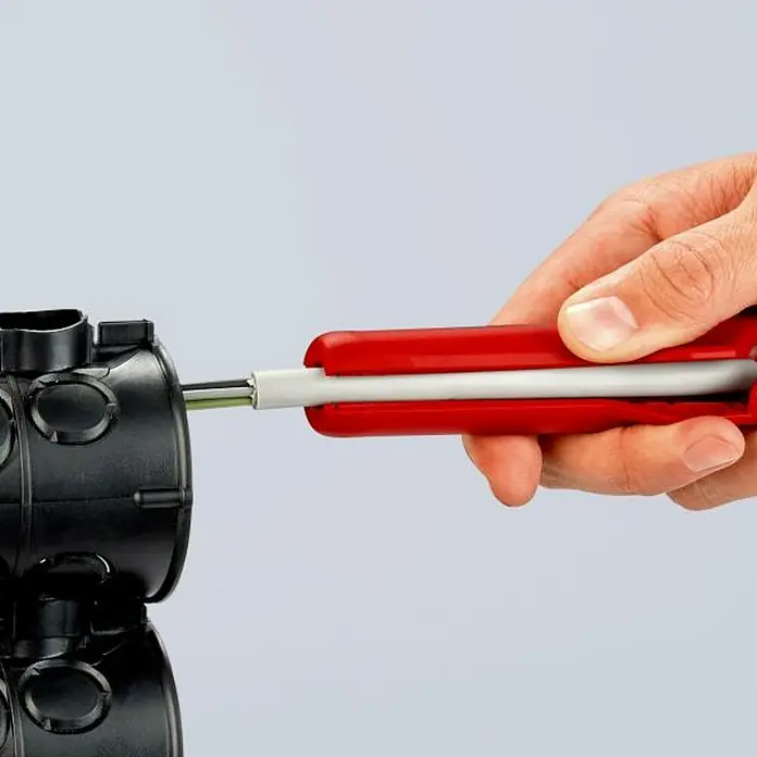 Knipex Pelacable - Largo: 17,5 cm - Detalle de unos alicates pelacables en acción, colocando un cable en una caja de conexiones negra, la herramienta tiene mangos rojos.