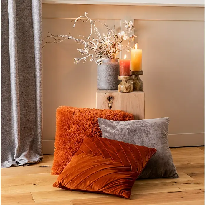 Kissen BodrumRostrot, 45 x 45 cm, 100 % Polyester - Dekoratives Stillleben mit Zierkissen in Orange, Rost und Grau, arrangiert mit Kerzen und Zweigen in einer grauen Vase auf einem Holzhocker.