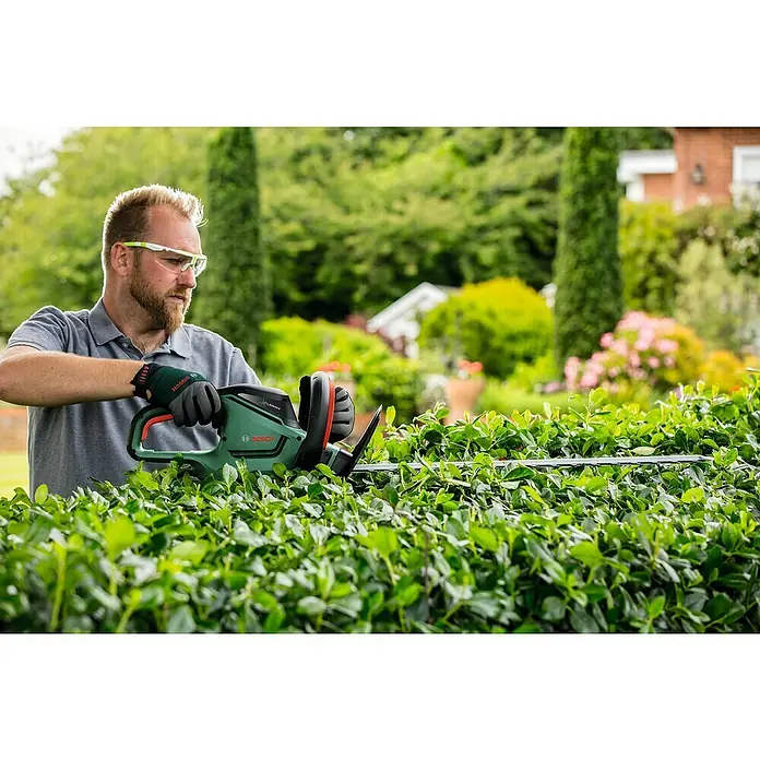 Bosch Elektro-Heckenschere UniversalHedgeCut 60 - 480 W, Schnittlänge: 60 cm - Ein Mann, der mit einer Bosch Heckenschere eine Hecke schneidet. Die Heckenschere ist grün und schwarz und hat eine lange Klinge.