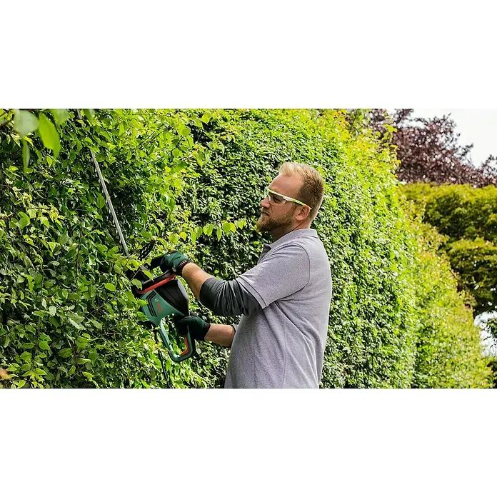 Bosch Elektro-Heckenschere AdvancedHedgeCut 70 - 500 W, Schnittlänge: 70 cm - Ein Mann trimmt eine Hecke mit einer Bosch Heckenschere, ausgestattet mit einer langen Klinge und grünen Akzenten.