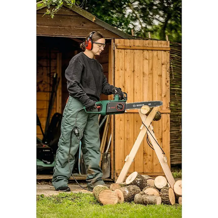 Bosch Elektro-Kettensäge UniversalChain 40 - 1.800 W, Schwertlänge: 40 cm - Eine Frau benutzt eine grüne Bosch Kettensäge, um Holz vor einer Holzhütte zu schneiden, und trägt dabei Schutzausrüstung.