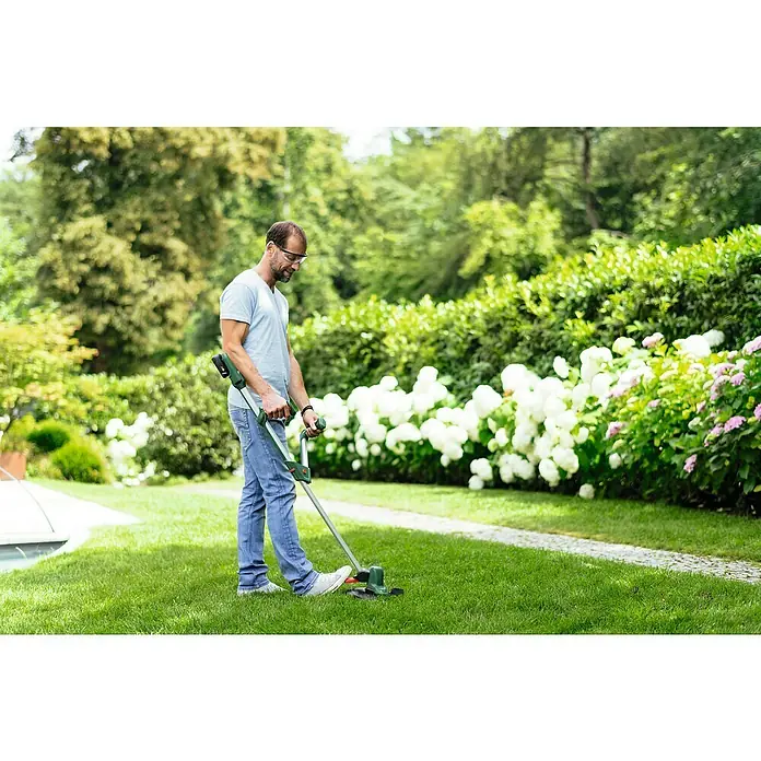 Bosch Power for All 18V Akku-Rasentrimmer UniversalGrassCut 18V-26 - 18 V, 2,5 Ah, 1 Akku - Ein Mann trimmt den Rasen mit einem grünen Rasentrimmer von Bosch, umgeben von blühenden Sträuchern, für einen gepflegten Garten.