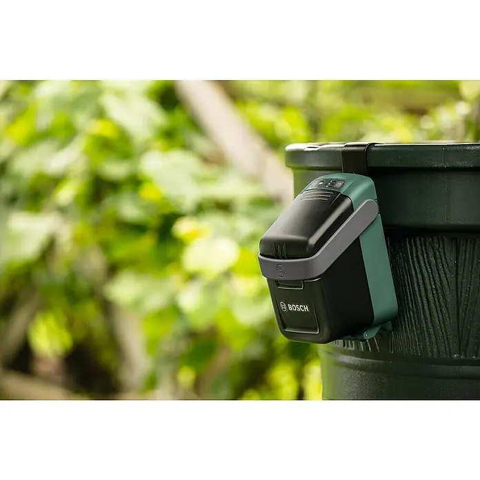 Bosch Power for All 18V Wandhalterung - Passend für: Bosch Akku-Regenwasserpumpe GardenPump 18 - Nahaufnahme des Bosch Gartenbewässerungssystems, das an einem dunklen Behälter befestigt ist, mit üppigem Grün im Hintergrund.