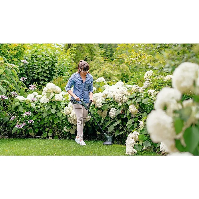 Bosch Power for All 18V Akku-Rasentrimmer EasyGrassCut 18V-26 - 18 V, 2,5 Ah, Ohne Akku - Eine Frau trimmt Gras mit einem Rasentrimmer. Der Rasentrimmer ist grün und hat einen Akku. Das Gras ist grün und der Hintergrund ist ein Garten.