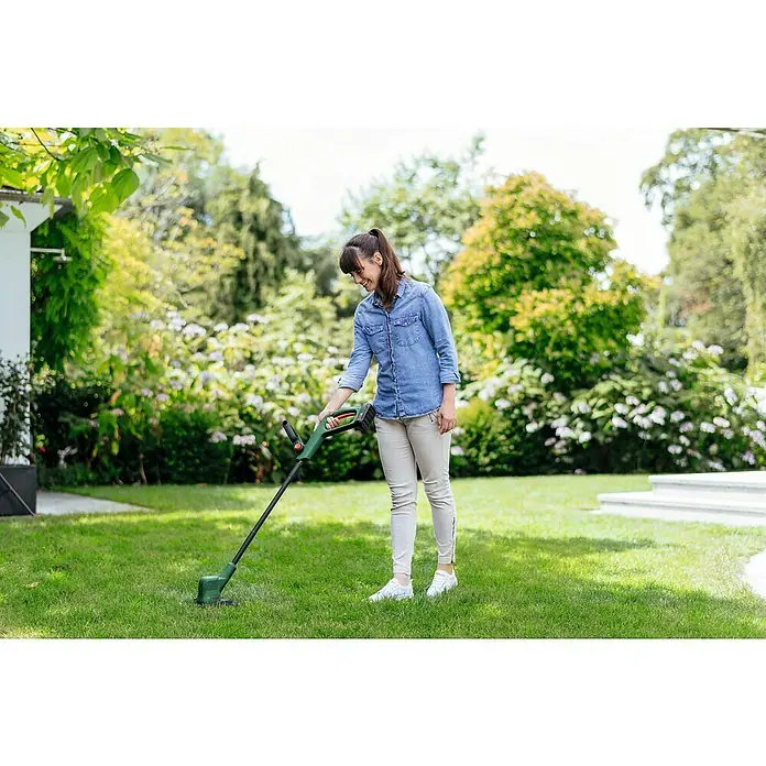 Bosch Power for All 18V Akku-Rasentrimmer EasyGrassCut 18V-260 - 18 V, 2 Ah, 2 Akkus - Eine Frau benutzt einen Rasentrimmer von Bosch im Garten, um Rasen zu schneiden. Das Gerät ist grün und schwarz, der Garten gepflegt.