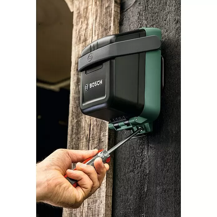 Bosch Power for All 18V Wandhalterung - Passend für: Bosch Akku-Regenwasserpumpe GardenPump 18 - Dunkelgrau-grüne Bosch Smart Watering Steuerung wird mit Schraubendreher an Holzwand montiert.