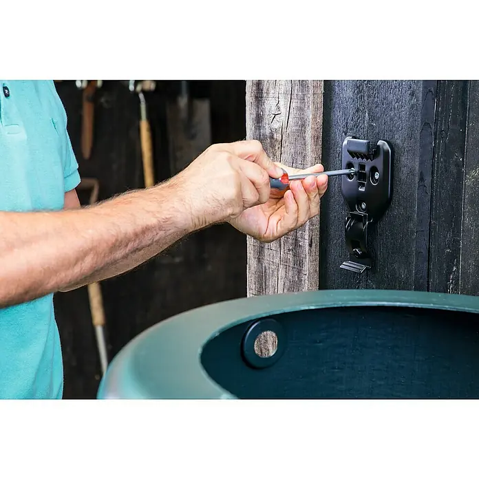 Bosch Power for All 18V Wandhalterung - Passend für: Bosch Akku-Regenwasserpumpe GardenPump 18 - Eine Person befestigt eine schwarze Halterung mit Schrauben an einer Holzwand. Fokus auf die Halterung und den Installationsprozess.