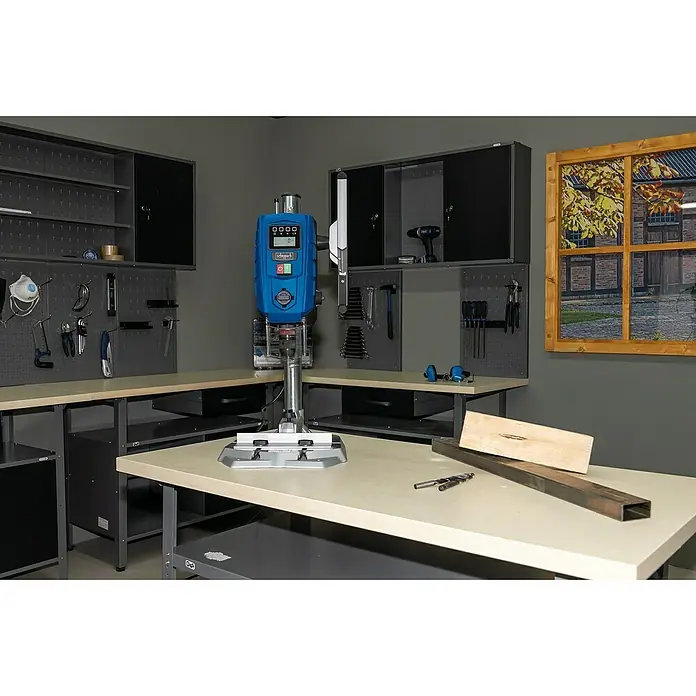 Scheppach Tischbohrmaschine DP 60 - 710 W, Leerlaufdrehzahl: 0 U/min - 2.600 U/min, Spannweite Bohrfutter: 1,5 mm - 13 mm - Eine Scheppach Tischbohrmaschine im Werkstattbereich, umgeben von Werkzeugen, Werkbänken und einem simulierten Fensterblick.