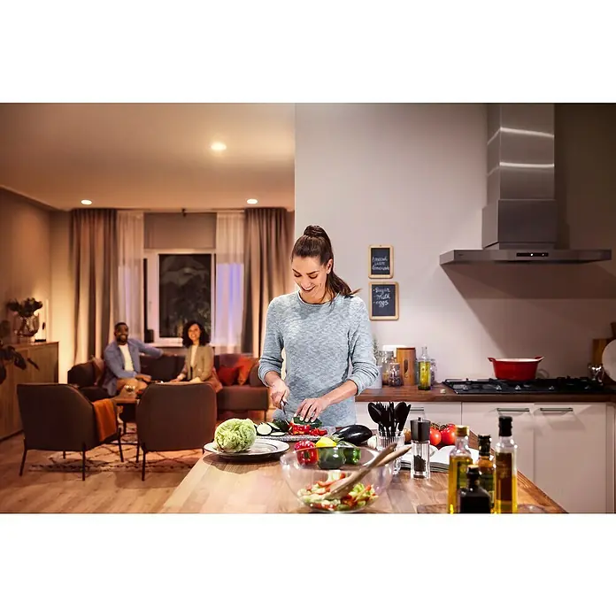 Philips Hue Smart-LED-Lampen Set White & Color AmbianceE27, Dimmbar, Warmweiß, 806 lm - 1.055 lm, 9 W - Eine Frau bereitet in einer modern ausgestatteten Küche mit Philips Hue Beleuchtung einen Salat zu, während sich andere im Wohnbereich entspannen.