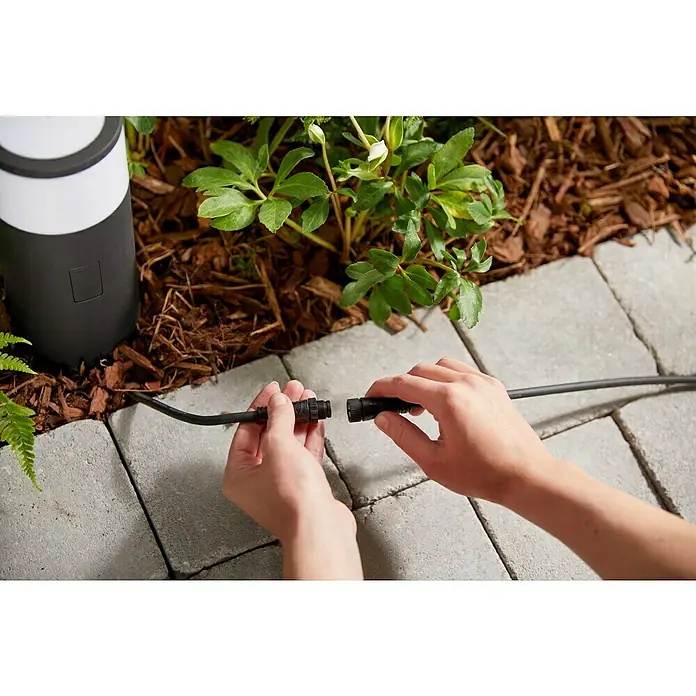 Philips Hue Verlängerungskabel OutdoorSchwarz, 250 cm - Eine Person verbindet ein schwarzes Kabel mit wasserdichten Anschlüssen im Garten, neben einer Gartenlampe.