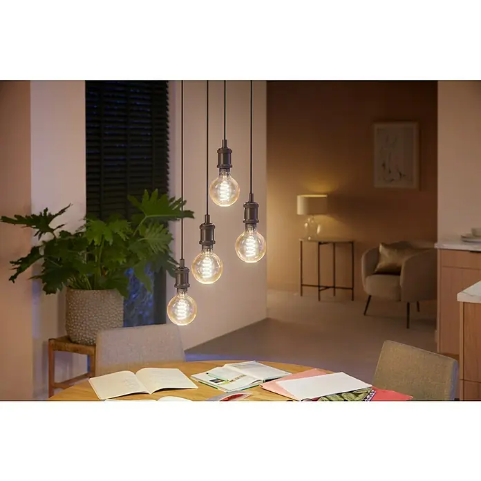 Philips Hue Bombilla LED inteligente White Ambiance Filament - E27, Intensidad regulable, Blanco cálido, 550 lm, 7 W, Denominación de la lámpara: G93, 1 ud. - Primer plano de una lámpara colgante decorativa con cuatro bombillas Edison colgando sobre una mesa de madera en una habitación iluminada cálidamente.