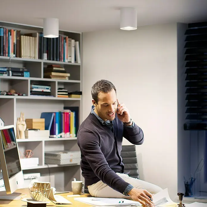 Philips Hue LED-Deckenstrahler Pillar - 5 W, L x B x H: 17,8 x 13,4 x 14,3 cm, Weiß, Neutralweiß - Mann sitzt auf Schreibtisch und telefoniert in einem Büro, das von zwei Deckenleuchten beleuchtet wird.