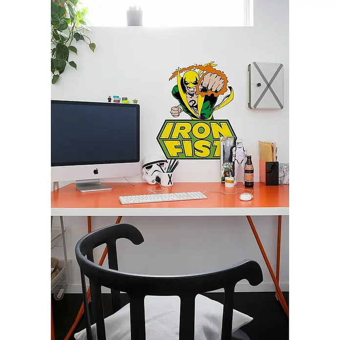 Komar Marvel Wandtattoo Iron Fist Comic - Bunt, 50 x 70 cm, 1 -tlg. - Schreibtisch mit orangefarbener Tischplatte, Computermonitor, Tastatur, Dekorationen und einem schwarzen Stuhl vor einer Wand mit Comic-Motiven.