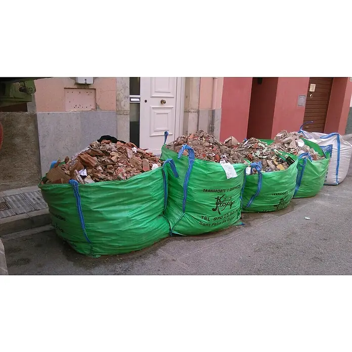 BHS Cataluña Big Bag para escombros zona Girona - 80 x 80 x 90 cm, Peso máximo admitido: 1.000 kg - Cuatro sacos grandes verdes llenos de escombros de construcción están sentados frente a un edificio listos para ser recogidos. Tienen el logo de una empresa de transportes.