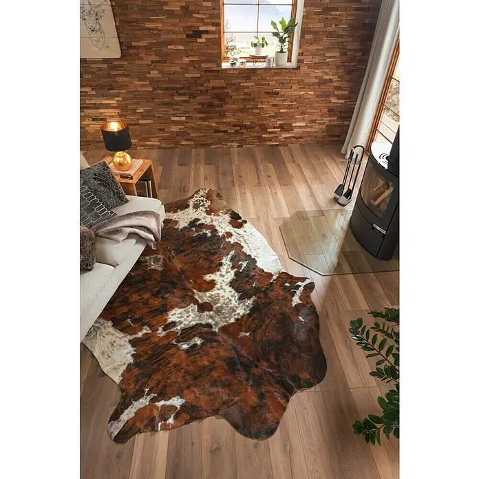 Esbeco Kuhfell - Exotisch Dunkel, 100 % Echtfell - Ein Kuhfellteppich in Braun- und Weißtönen liegt auf einem Holzboden und verleiht dem Raum eine rustikale Note.