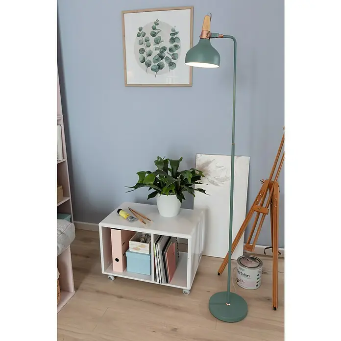 SCHÖNER WOHNEN-Farbe Wandfarbe Designfarbe - Klares Eisblau, Matt, 2,5 l, Konservierungsmittelfrei - Eine grüne Stehlampe mit hölzernen Akzenten beleuchtet eine Szene mit einem Tisch, Pflanzen und einem Kunststaffelei.