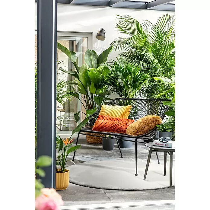 Alfombra de pelo Happy - Plateado, Diámetro: 120 cm, 100 % poliéster (pelo / felpa) - Banco de exterior de metal negro con tres cojines coloridos en un patio lleno de plantas.