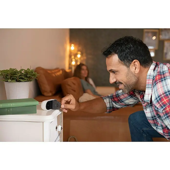Abus Überwachungskamera Privacy - Grau, Weiß - Ein Mann, der eine kabellose Blink-Kamera auf einem Nachttisch mit Pflanze platziert, während eine Frau auf dem Sofa sitzt.