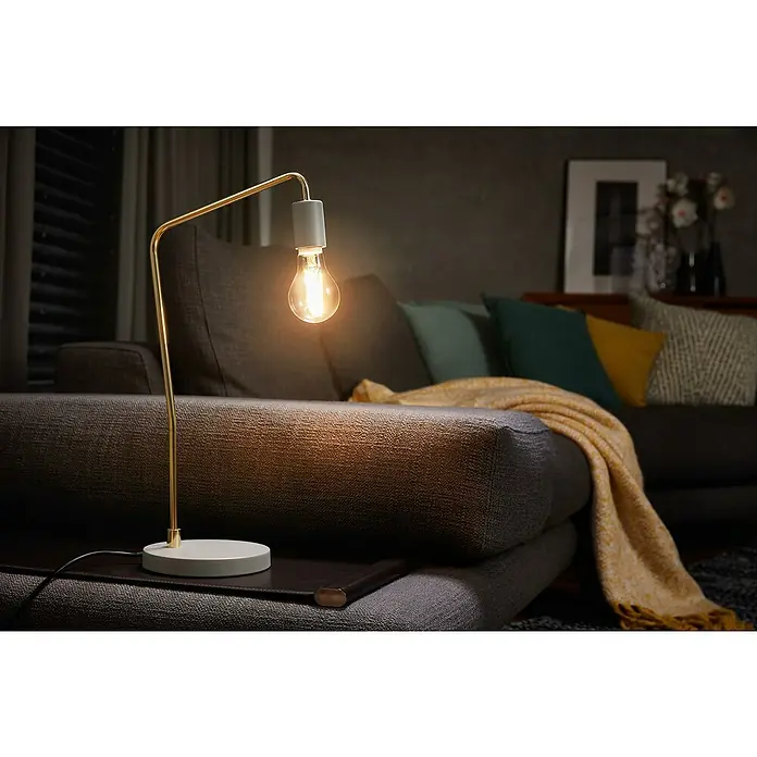 Voltolux Bombilla LED con filamento Classic A - E27, No regulable, Blanco cálido, 470 lm, 4 W - Primer plano de una elegante lámpara de mesa con bombilla Edison, que emite una luz cálida sobre un sofá moderno.