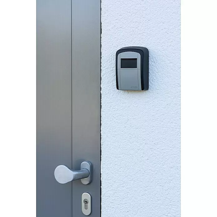 Stabilit Schlüsselbox CBO 704 - L x B x H: 8,8 x 3,9 x 12 cm, Schwarz/Silber - Grau-schwarzer ABUS Schlüsselsafe an weißer Wand neben grauer Tür mit silbernem Griff.