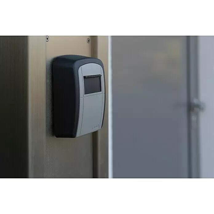 Stabilit Schlüsselbox CBO 704 - L x B x H: 8,8 x 3,9 x 12 cm, Schwarz/Silber - Schwarzer und grauer Schlüsseltresor, montiert an einer gebürsteten Metallwand neben einer unscharfen Tür.