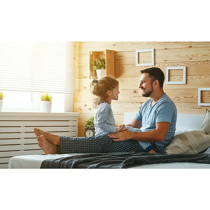 Blockhausprofil - Fichte/Tanne, L x B: 3.000 x 121 mm - Vater und Tochter sitzen zusammen auf dem Bett, wobei die Tochter auf dem Schoß des Vaters sitzt. Sie lachen und haben Spaß.