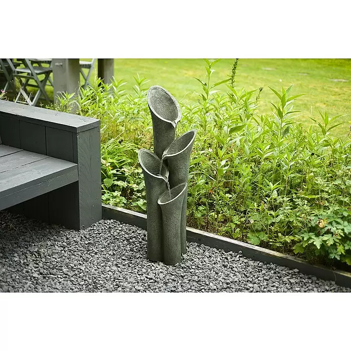 Ubbink AcquaArte Gartenbrunnen Montreux - 23,5 x 25 x 86 cm, Kunststoff, Grau - Grauer Gartenbrunnen mit mehreren Stufen, aus dem Wasser fließt, auf Kies neben einer Bank.